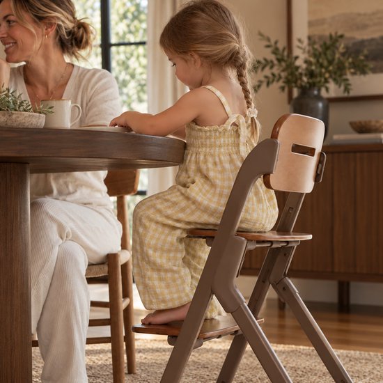 Kind in geel-witte ruitjes jumpsuit zittend op een donkerbruine Deryan houten kinderstoel aan de eettafel.