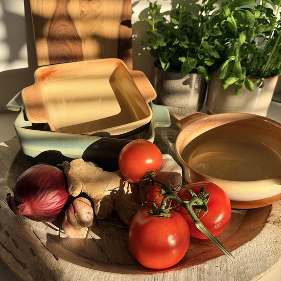 Vierkante terracotta ovenschaal met blauwgroene buitenkant, omringd door verse tomaten, ui, knoflook en gember op een dienblad.