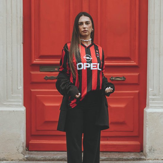 Model draagt een rood-zwart gestreept AC Milan retroshirt met OPEL-logo voor een rode deur.