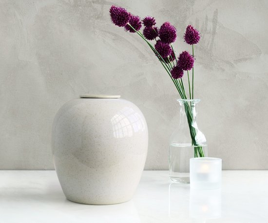 Witte keramische urn met spikkels, naast een glazen vaas met paarse bloemen en een kaarsenhouder.