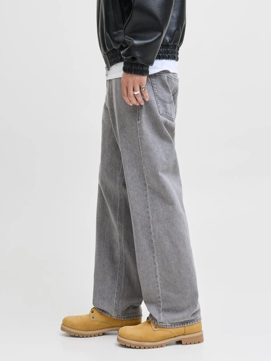 Man van de zijkant in wijde grijze jeans, zwarte jas en gele laarzen, staand op een lichte achtergrond.