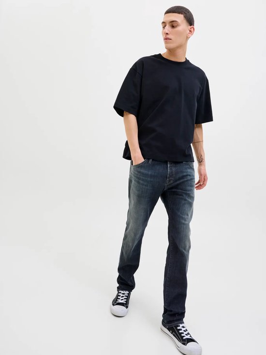 Man met donkere jeans en zwart oversized T-shirt, staand met handen in de zakken, op de voorgrond.