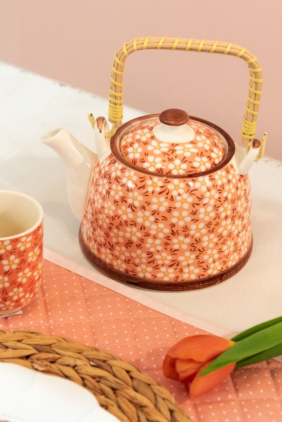 Koraalkleurige theepot met een vrolijk bloemenpatroon in wit en geel, met een rieten handvat.