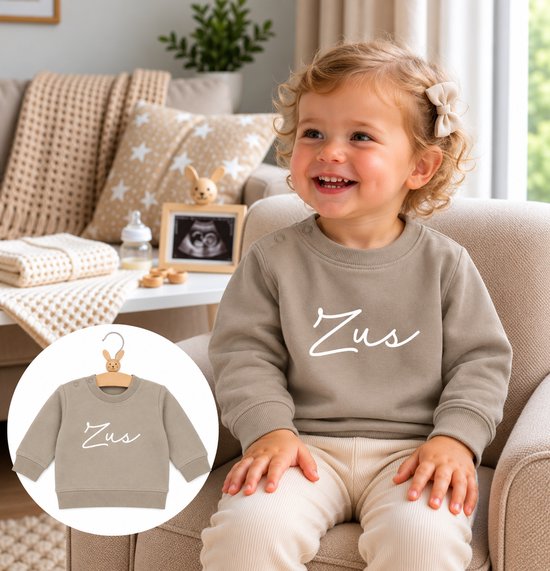 Beige kindertrui met het woord 'Zus' in witte sierletters, gedragen door een lachend kind met een strikje.