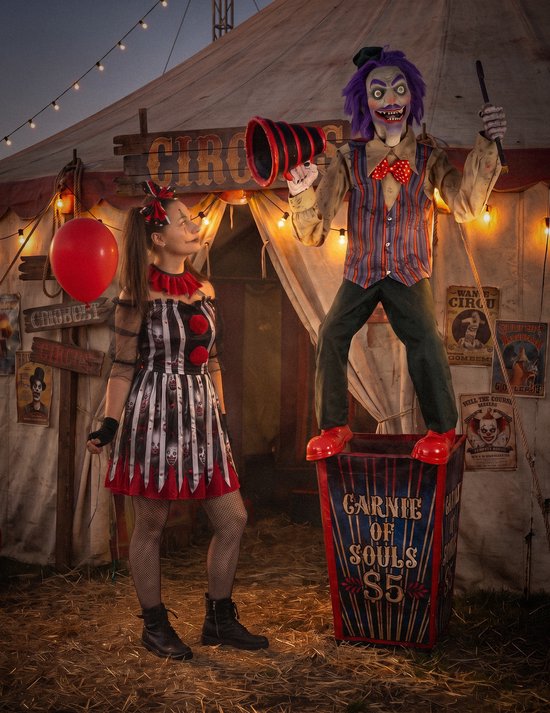 Vrouw in clownskostuum naast een enge pop op een 'Carnie of Souls'-doos met paars haar en megafoon voor circustent.