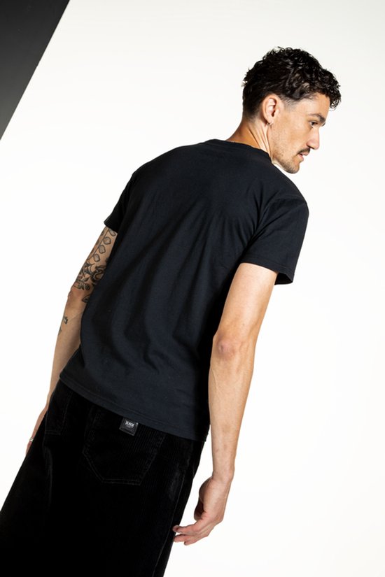 Model met getatoeëerde arm draagt een zwart T-shirt en een zwarte broek van corduroy.