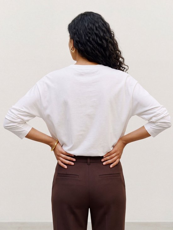 Achterzijde van een model met een wit shirt met lange mouwen en een donkerbruine pantalon.