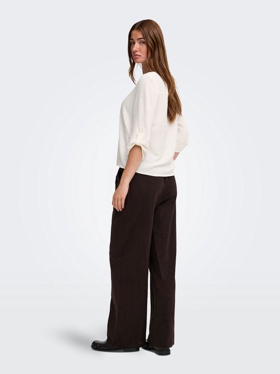 Zijaanzicht van een model in witte blouse met kreuktextuur en donkerbruine broek.