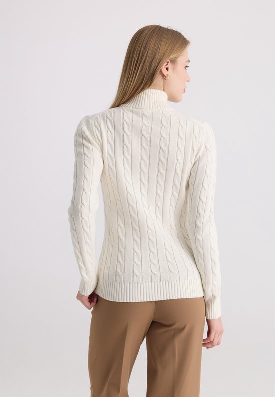 Zijaanzicht van een model in een ecru kabelgebreide trui met halve rits en een bruine broek.