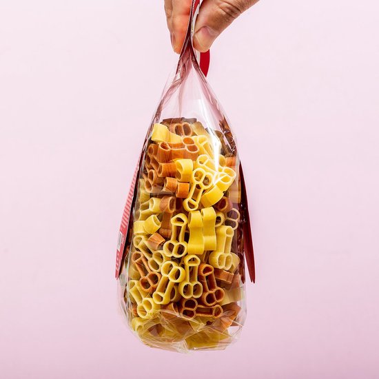Transparante zak met droge pasta in de vorm van penissen, in geel en bruin, tegen een zachte roze achtergrond.