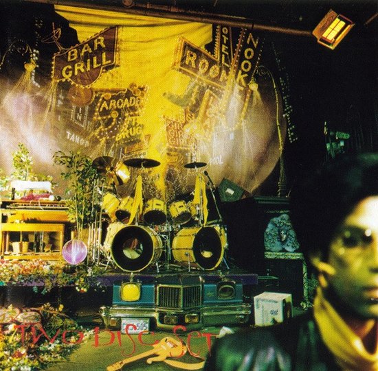 Prince op de voorgrond voor een drumstel op een podium, met neonborden en de tekst 'TWO DISC SET'.