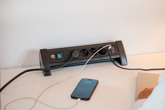 Zwarte stekkerdoos met 4 stopcontacten en 2 USB-poorten, aangesloten op een smartphone en een laptop, op een bureau.
