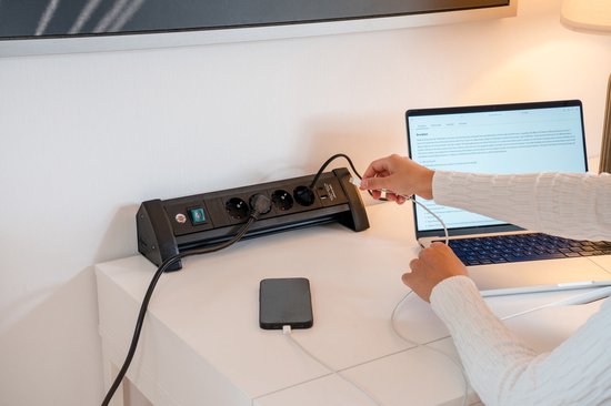Zwarte stekkerdoos met 4 stopcontacten en 2 USB-poorten, aangesloten op een smartphone en een laptop, op een bureau.