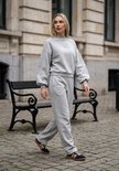 Model in grijze joggingbroek en sweater met pofmouwen. Een logo is subtiel op de borst van de sweater geborduurd.
