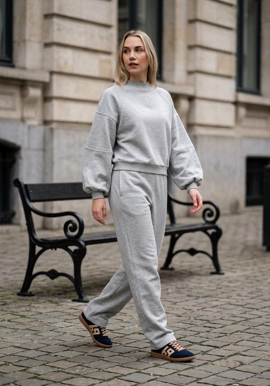 Model in grijze joggingbroek en sweater met pofmouwen. Een logo is subtiel op de borst van de sweater geborduurd.