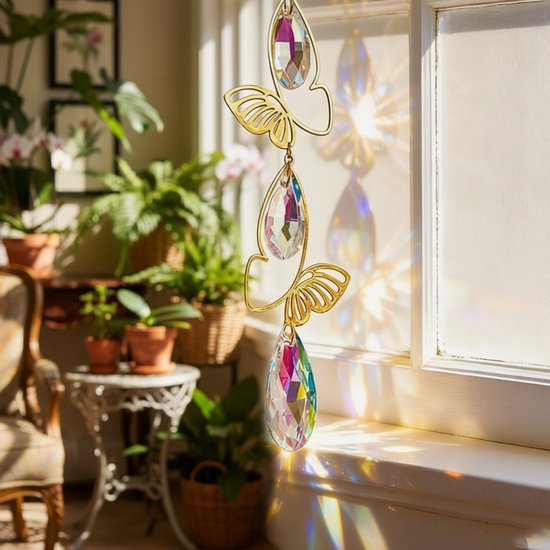 Gouden hanger met vlinders en druppelvormige kristallen, aan een raam met zonlicht.
