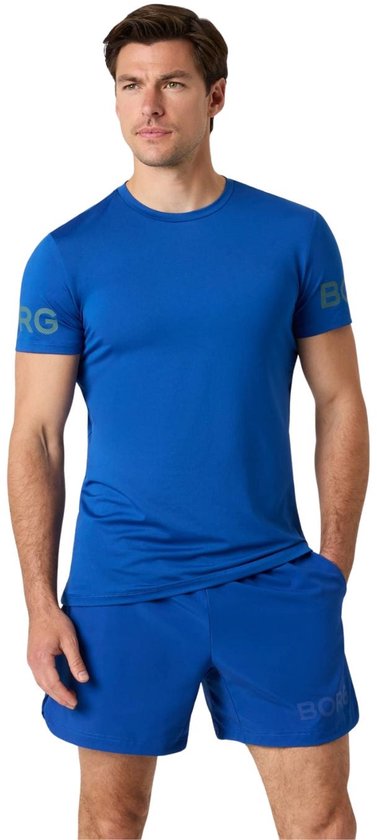 Model draagt een blauwe Björn Borg sportset: een t-shirt met korte mouw en shorts. Het t-shirt heeft een logo op de mouw en de shorts hebben een logo op de voorkant.