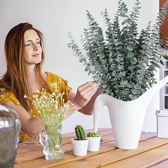 Vrouw met lang rood haar in een gele jurk, zet eucalyptus takken in een witte kan. Kleine cactussen en bloemetjes staan op tafel.