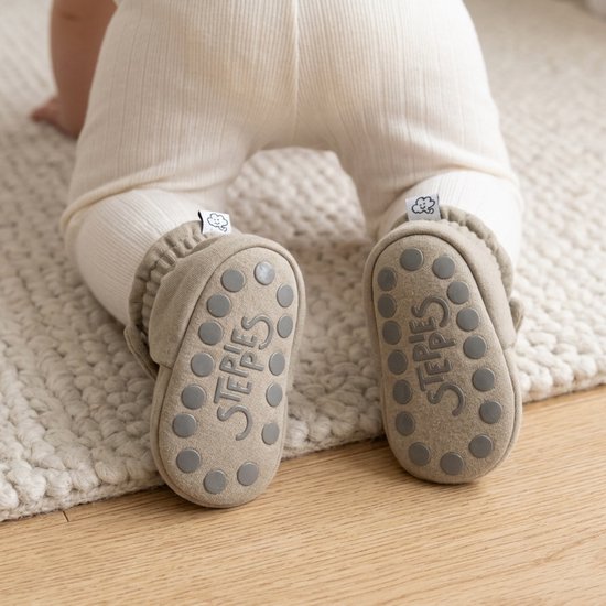 Baby op handen en knieën draagt taupe slofjes met antislip-zolen waarop de tekst Steppies staat.