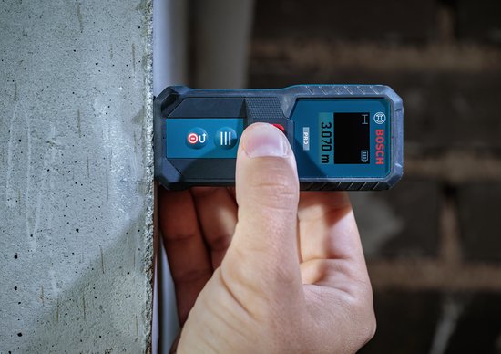 Hand houdt een blauwe Bosch laser-afstandsmeter vast tegen een muur met het display op 3,070 m.