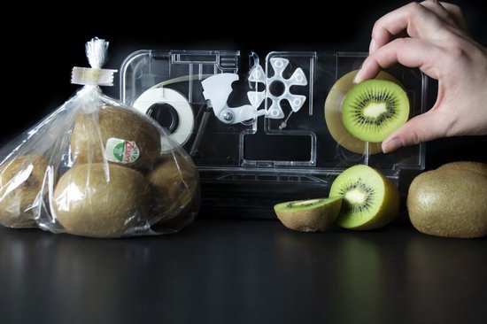 Een zak kiwi's en een hand die een kiwiplakje bij een transparant sealapparaat houdt.