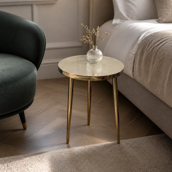 Table d'appoint ronde dorée avec un plateau poli et des pieds fins, à côté d'un fauteuil vert et d'un lit.
