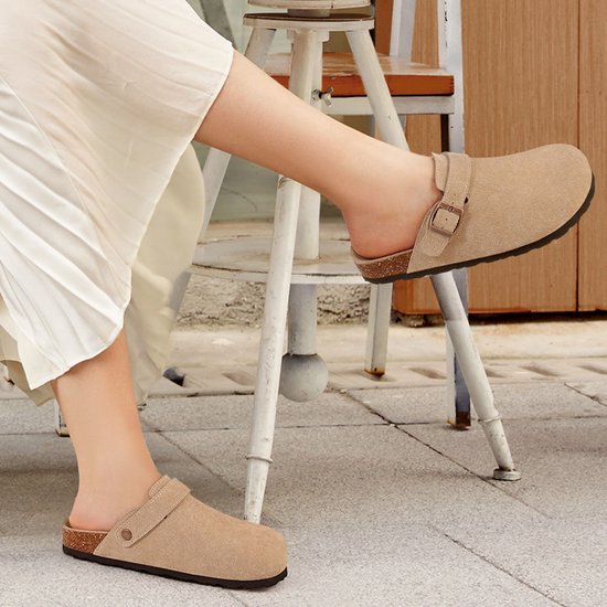 Een vrouw draagt beige suède klompjes met een gesp en een verstelbare band over de wreef.