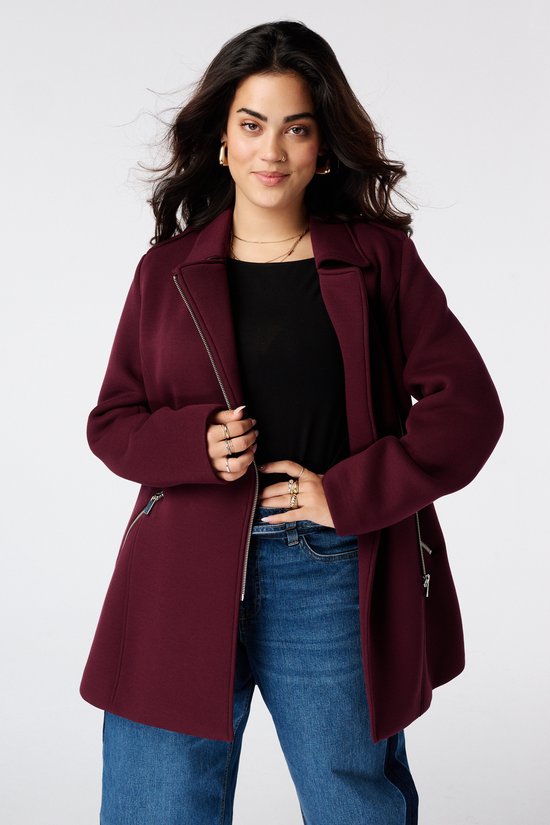 Model met bordeauxrode jas open, zwarte top en jeans, handen op de ritssluiting.