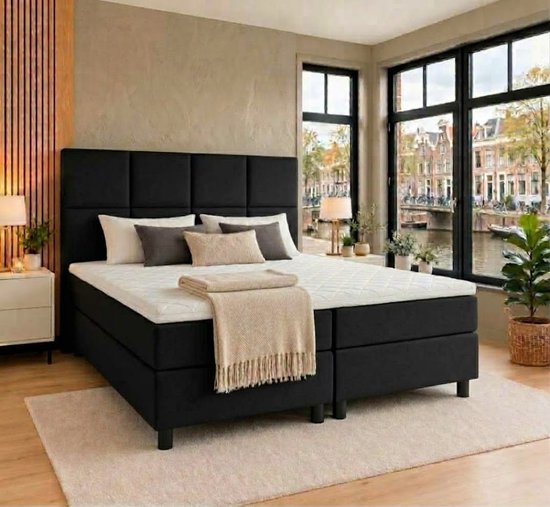 Zwarte boxspring met gestoffeerd hoofdbord, wit matras en beige plaid in een modern, licht interieur.