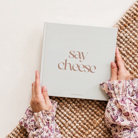 Handen in een bloemetjesblouse houden een beige 'say cheese' schoolfotoboek vast op een rieten ondergrond.