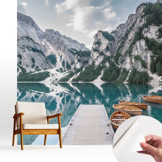 Bergpanorama fotobehang op een muur achter een fauteuil, met een hand die een stukje zelfklevend vliesbehang optilt.