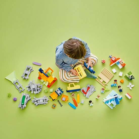 Kind van bovenaf gezien, zittend op een groene ondergrond, omringd door losse LEGO Duplo treinonderdelen.