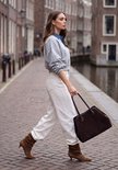 Model in grijze sweater met blauwe kraag, witte broek, bruine suède laarzen en donkere handtas in de stad.