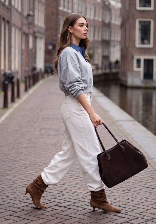 Model in grijze sweater met blauwe kraag, witte broek, bruine suède laarzen en donkere handtas in de stad.