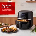 Zwarte heteluchtfriteuse met open mand vol friet. Naast de airfryer ligt een bord met steak, groente en friet. "Gezonder bakken Tot 90% minder vet, toch net zo krokant."