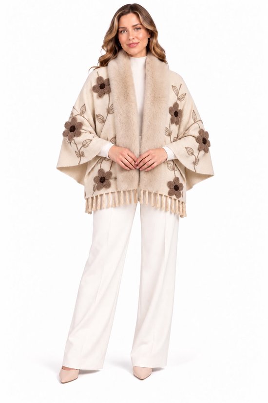 Vrouw in crèmekleurige poncho met imitatiebontkraag, bruine 3D-bloemen en franjes, gedragen over een witte coltrui en witte broek.