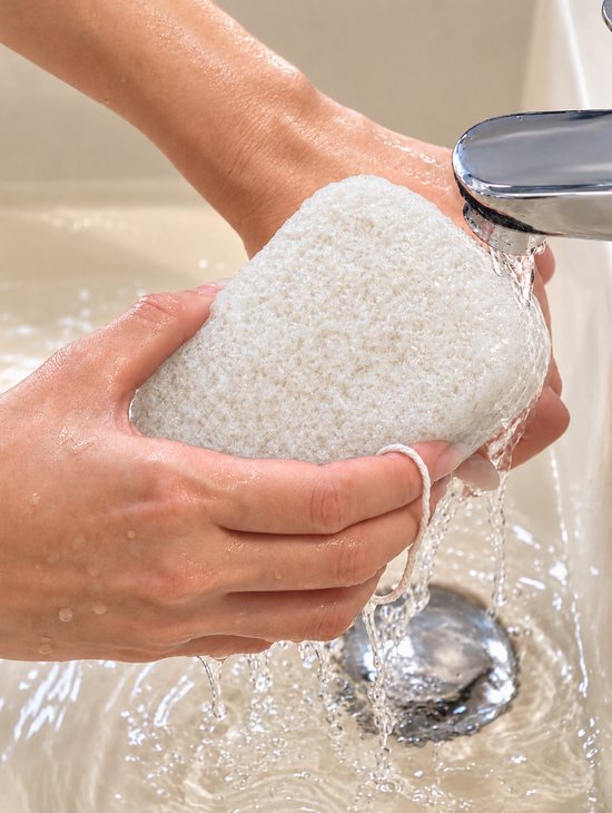 Handen houden een witte konjac-badspons vast terwijl deze onder een stromende kraan in een wastafel wordt afgespoeld.