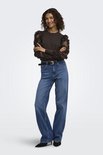 Model in bruine trui met doorschijnende mouwen met bloemapplicaties, gekruiste armen en jeans.