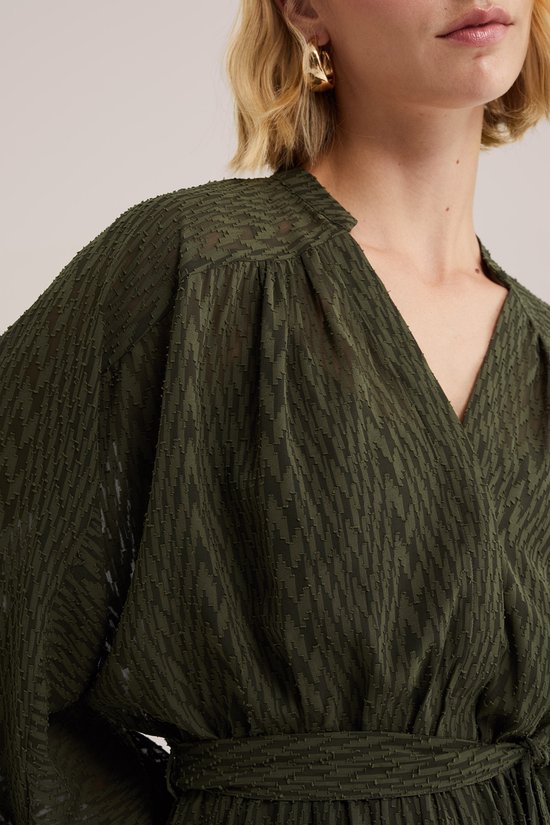 Close-up van een donkergroene dames jurk met gedetailleerde stof en V-hals. Met goudkleurige oorbel.
