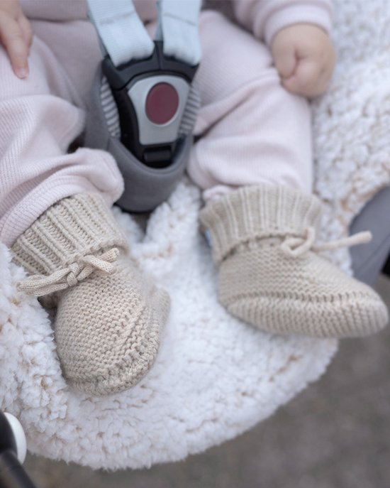 Een baby in een kinderwagen, met beige gebreide slofjes, een zachte deken en roze kleding.