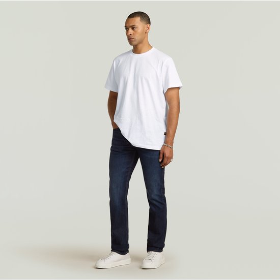Man in een witte T-shirt en donkerblauwe G-Star jeans, staand met een hand in de zak. Zijaanzicht.