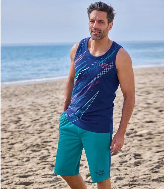 Man op strand in marineblauw sportshirt met blauwe grafische print en een petrolgroene short met zijkantstreep.
