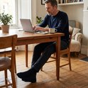 Man op donkerblauw pakbroek en sokken werkt op laptop.