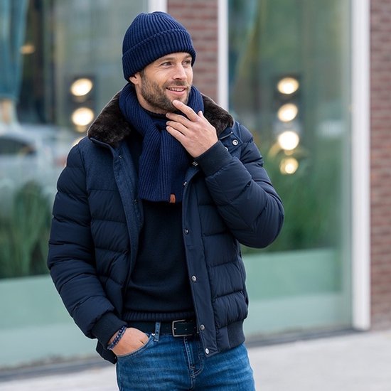 Homme portant une veste bleu marine avec col en fourrure, une écharpe, un bonnet, un pull bleu marine et un jean bleu. Ceinture bleu marine avec boucle carrée.