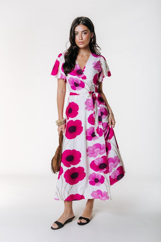 Model in een witte wikkelmaxi-jurk met grote roze bloemenprint en strikceintuur. Draagt zwarte sandalen en gouden armbanden.