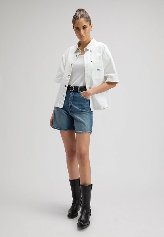 Een vrouw draagt een spijkerbroek met wijde pijpen, een witte tanktop, een witte korte mouwen top en zwarte cowboylaarzen.