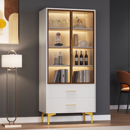 Witte vitrinekast met glazen deuren, gouden handgrepen, gouden poten en warme LED-verlichting. Gedecoreerd met boeken, flessen en beeldjes.