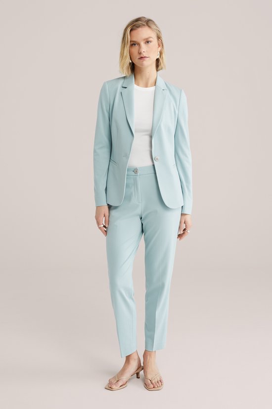 Vrouw in een lichtblauwe getailleerde blazer en bijpassende broek, staand met één hand in de zak.