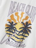 Detail van een witte T-shirt met een print van palmbomen en zonsondergang.
