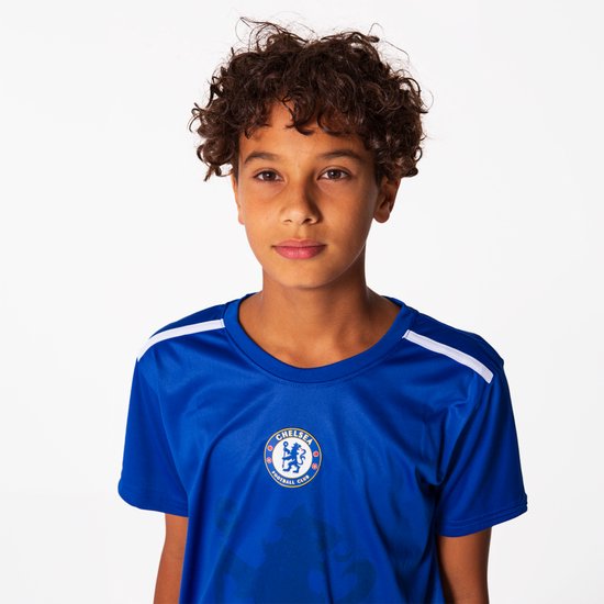 Jongen met krullend haar, poseren in een effen blauw Chelsea FC voetbalshirt met witte schouderstrepen.